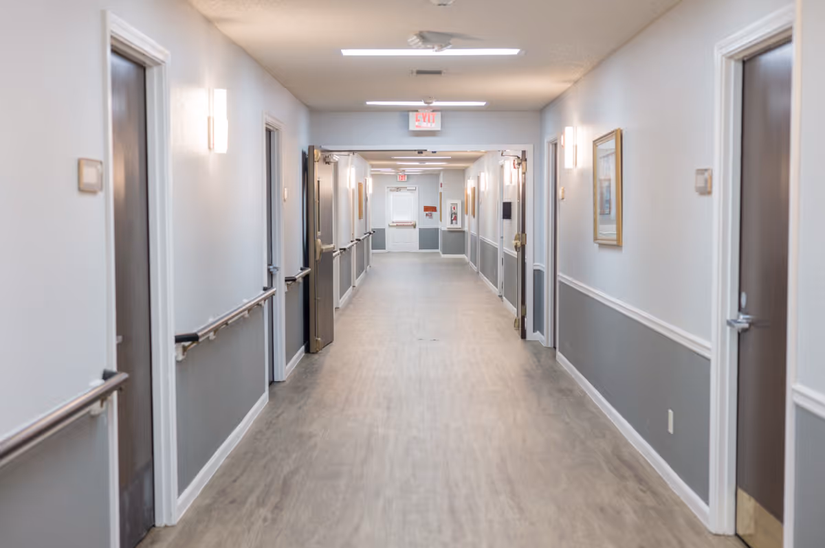 A long, clean hallway in a senior living facility with light gray walls, wooden doors on both sides, handrails along the walls, and overhead fluorescent lighting. There are framed pictures on the walls and an exit sign at the far end of the corridor.