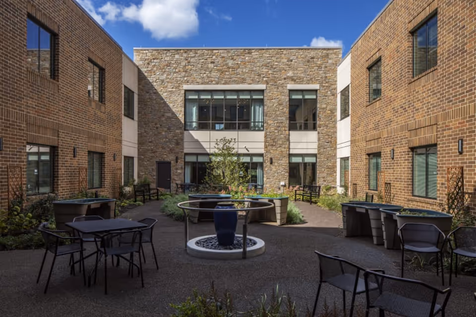 Outdoor courtyard area at Eagleview Landing featuring brick and stone building walls, several black metal tables and chairs, raised garden beds, a circular water fountain in the center, and some greenery under a partly cloudy blue sky.