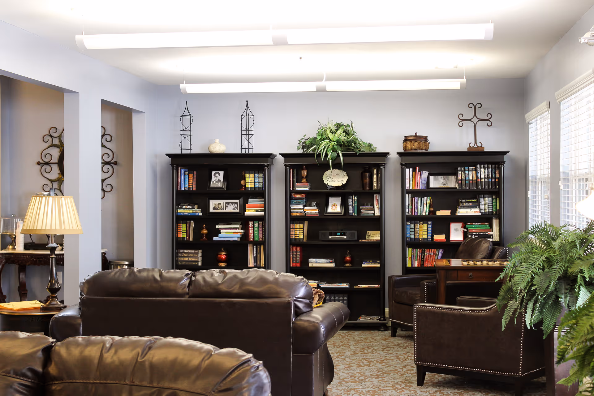 A communal living room with leather sofas, armchairs, bookshelves, lamps, and plants.