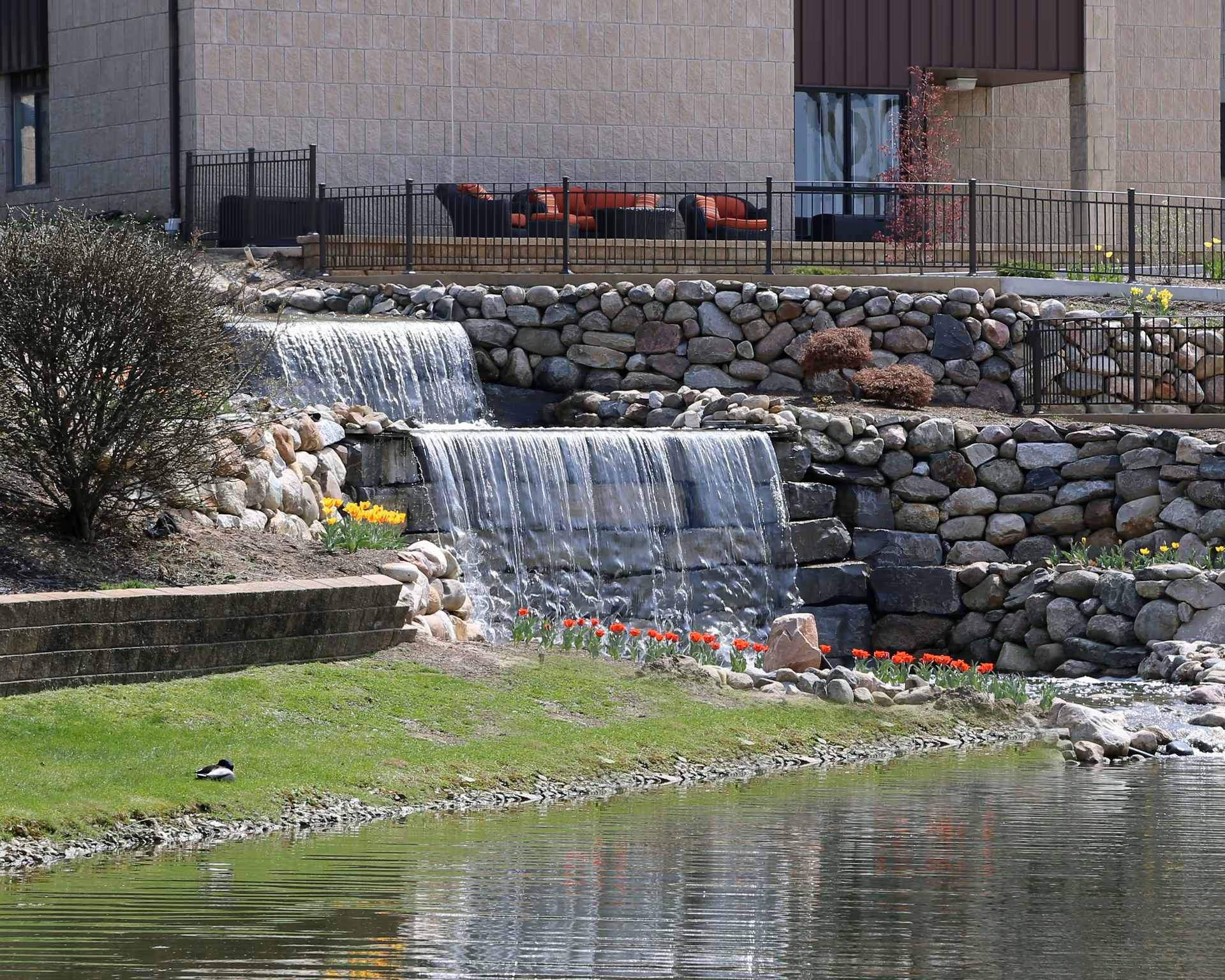 A landscaped outdoor area featuring a small cascading waterfall over stone walls into a pond, with red and yellow flowers planted along the water's edge. In the background, there is a building with a patio area furnished with outdoor seating.