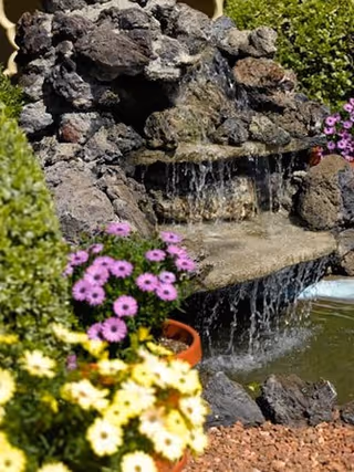 A small outdoor rock waterfall feature surrounded by green bushes and colorful flowers including purple and yellow blooms.