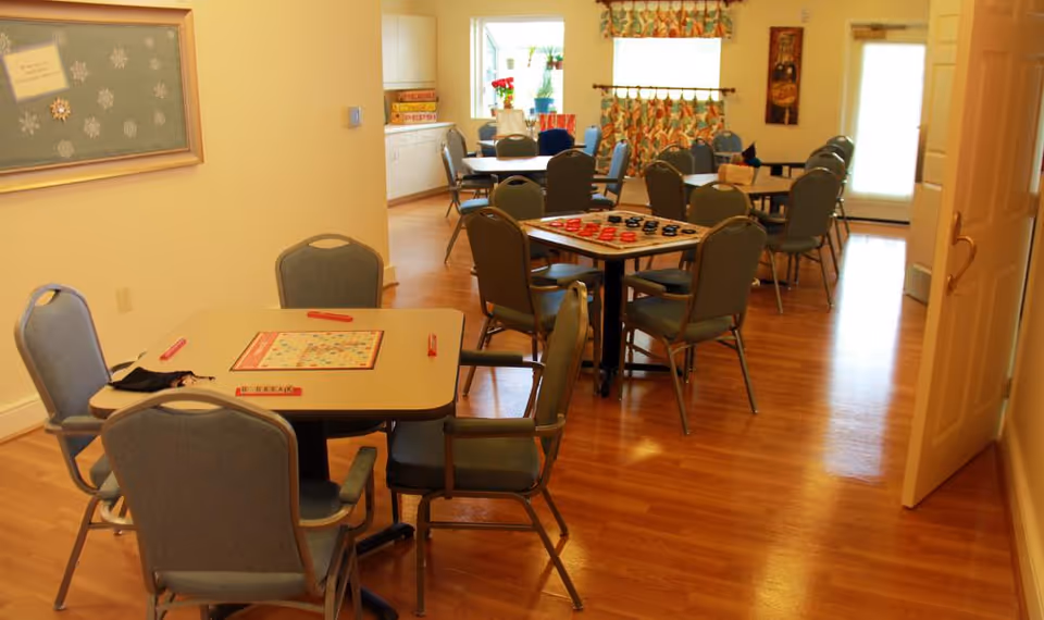 A well-lit room with multiple tables and chairs arranged for group activities. One table in the foreground has a Scrabble board game set up, while another table in the background has a checkers game. The room has wooden flooring, light-colored walls, a window with floral curtains, and a door on the right side.