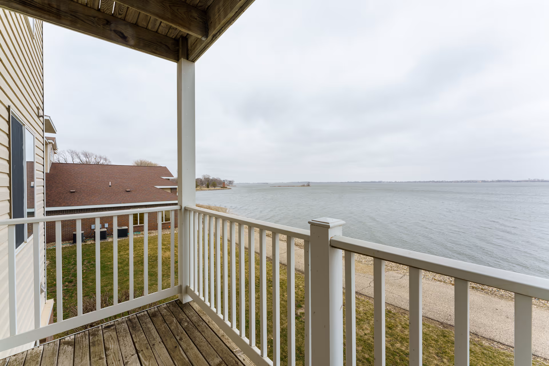 View from a wooden balcony with white railings overlooking a large body of water under a cloudy sky. A building with beige siding and a brown roof is visible to the left, along with a grassy area and a paved path near the water's edge.