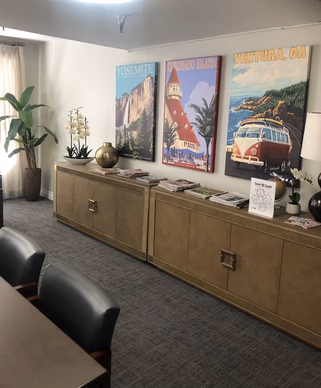 Common room interior with a long sideboard holding magazines, decorative vases and plants beneath three colorful California travel posters, with chairs and a table in the foreground.