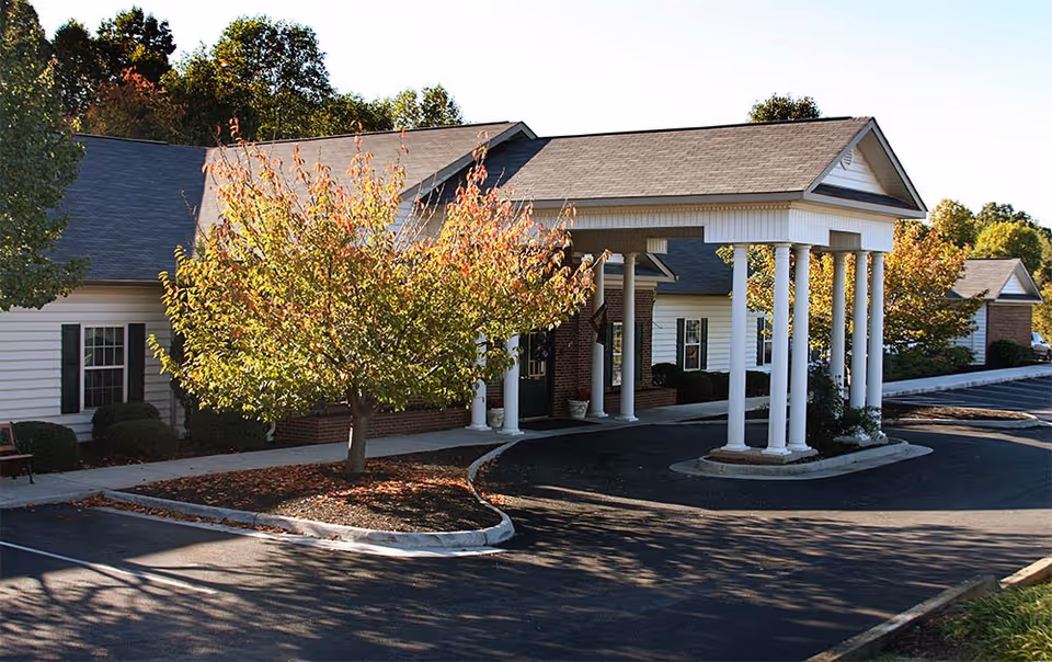 Exterior view of a single-story building with white siding and a gray roof, featuring a covered entrance supported by white columns. There is a tree with autumn-colored leaves in front of the building and a paved driveway and parking area surrounding it.