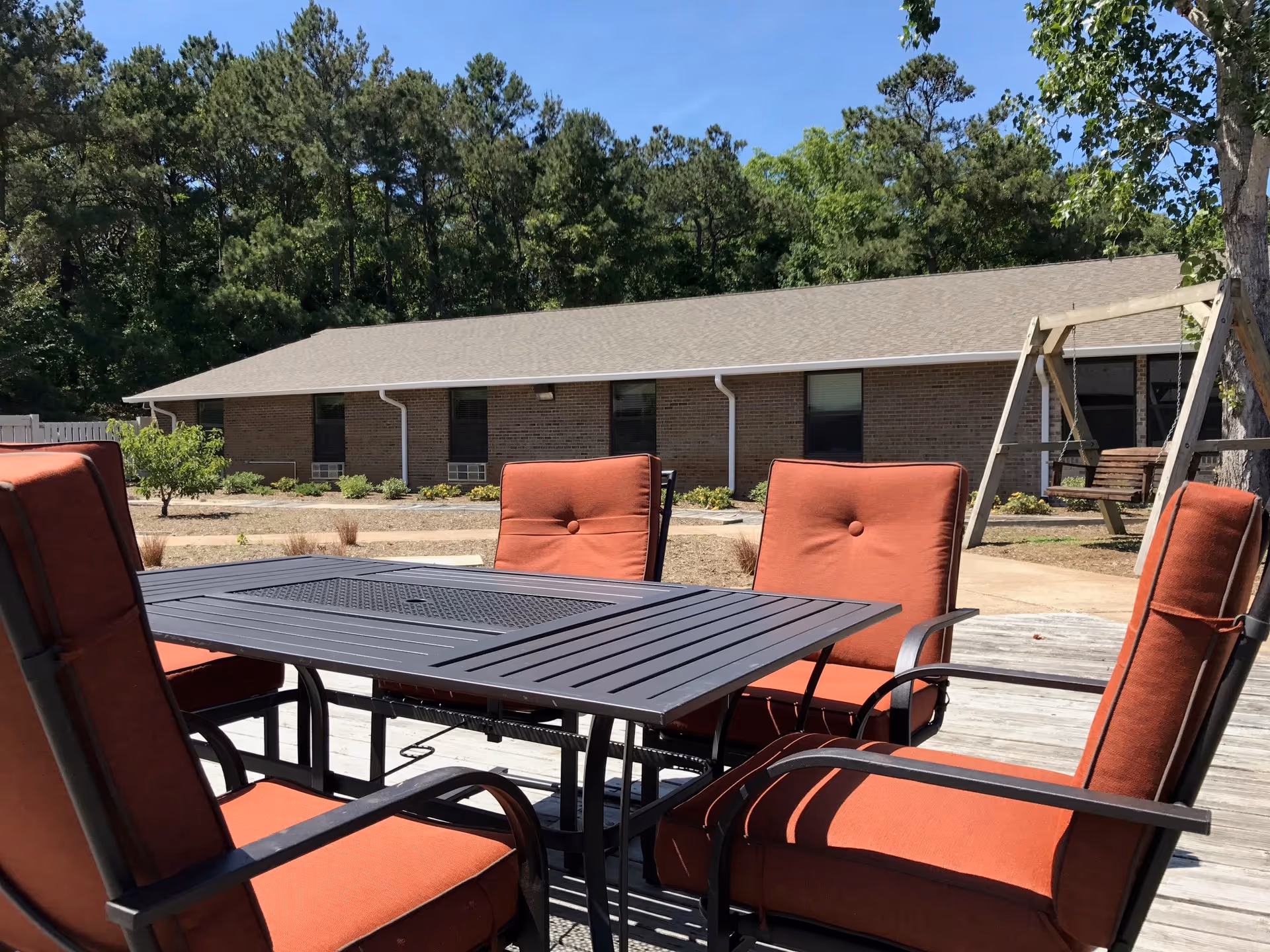 Patio table with red cushioned chairs on a wooden deck in front of a single-story brick building and trees.