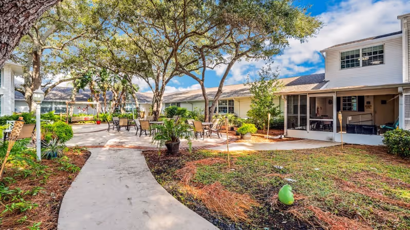 Outdoor courtyard area at The Pointe featuring a paved walkway, several tables and chairs under large trees, surrounded by greenery and shrubs, with a white building in the background under a partly cloudy sky.