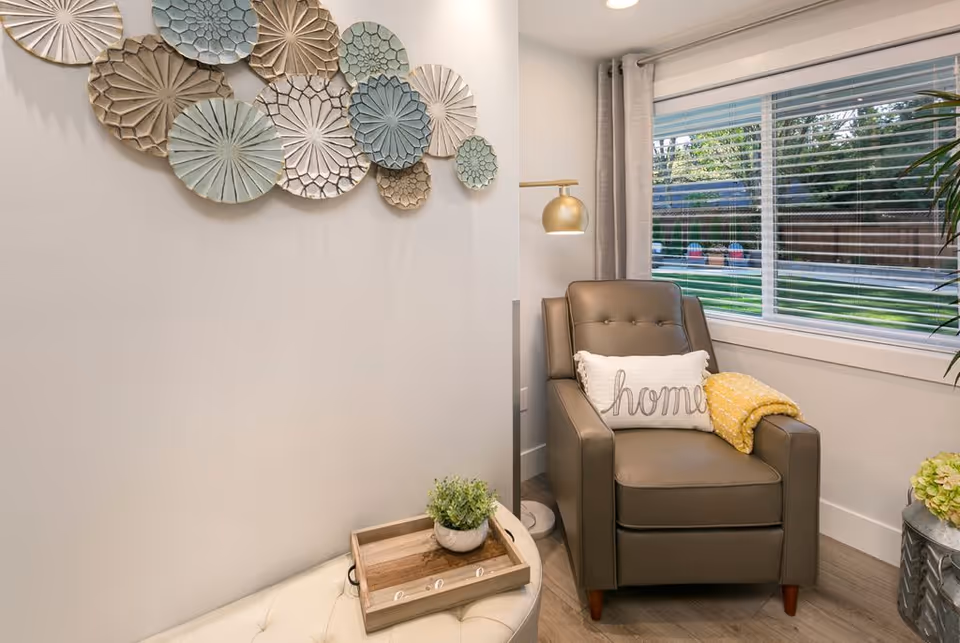 A cozy corner in a living room featuring a brown leather armchair with a white pillow that says 'home' and a yellow throw blanket. Next to the chair is a floor lamp with a gold shade. A window with blinds shows a green outdoor area with chairs. On the wall above a white cushioned bench is a decorative arrangement of circular, textured wall art in neutral tones. A small wooden tray with a potted plant sits on the bench.