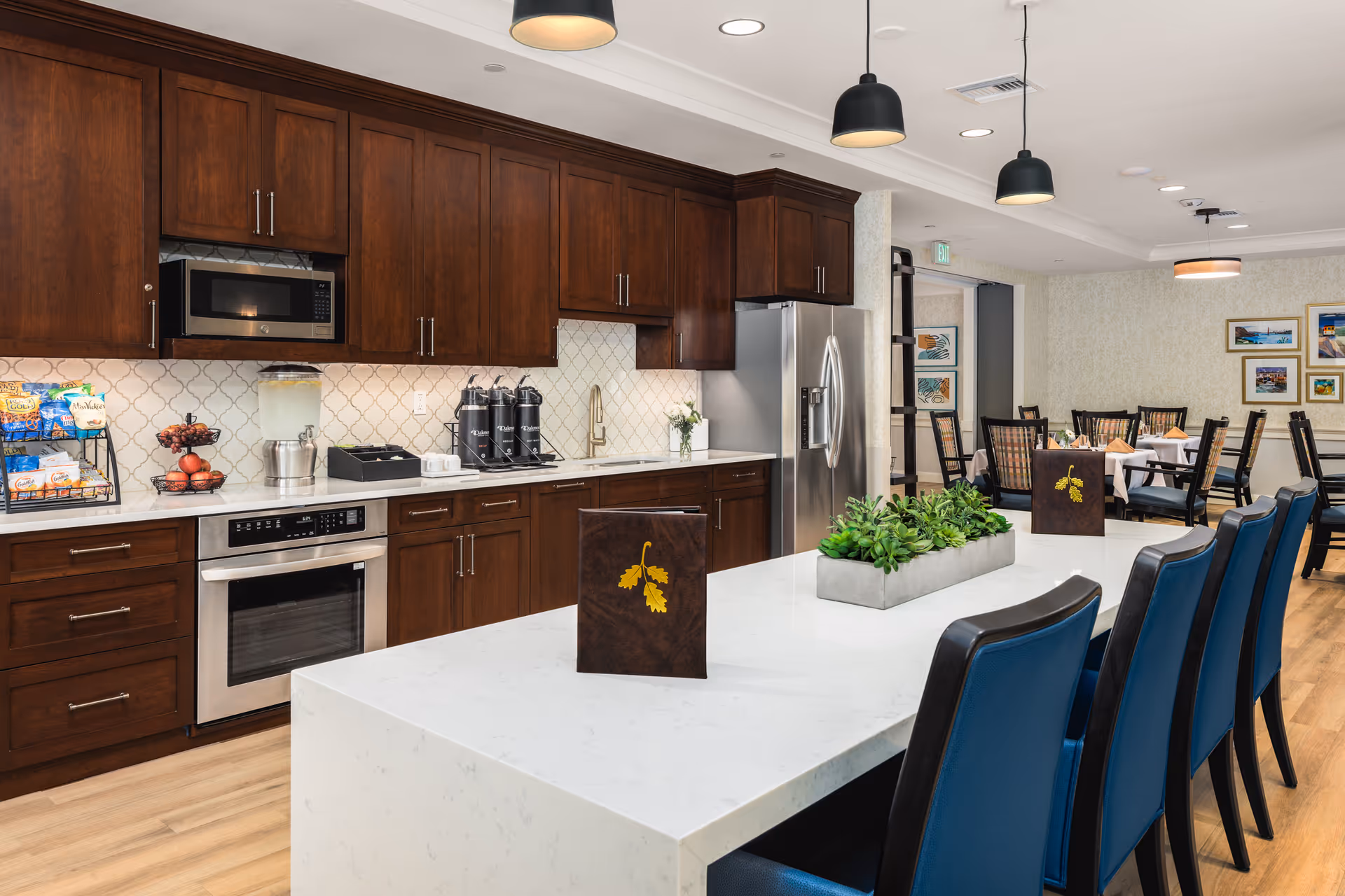 A modern kitchen and dining area in a senior living facility with dark wooden cabinets, stainless steel appliances, and a white marble island countertop with blue chairs. The dining area has tables with chairs and framed artwork on the walls.