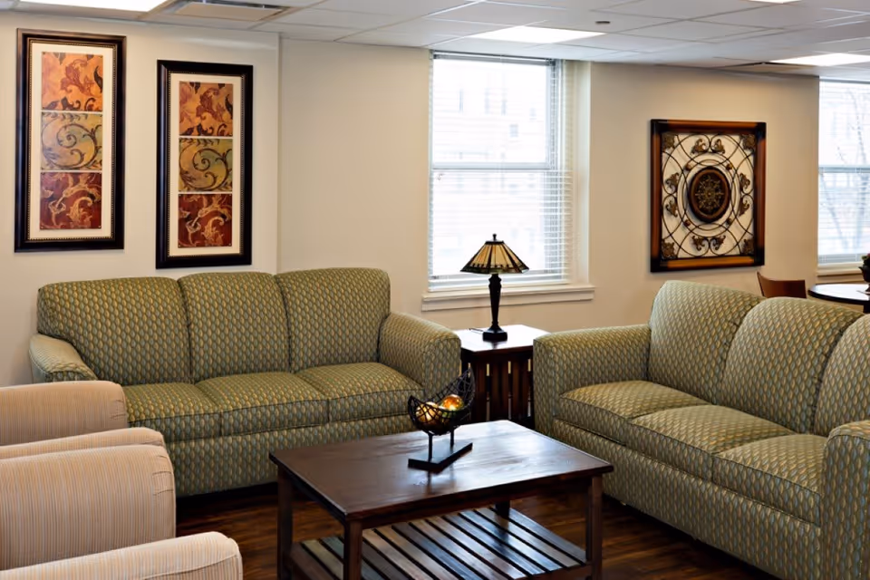 A seating area with two patterned green sofas, armchairs, a wooden coffee table, side table with a lamp, and framed wall art in a well-lit room.