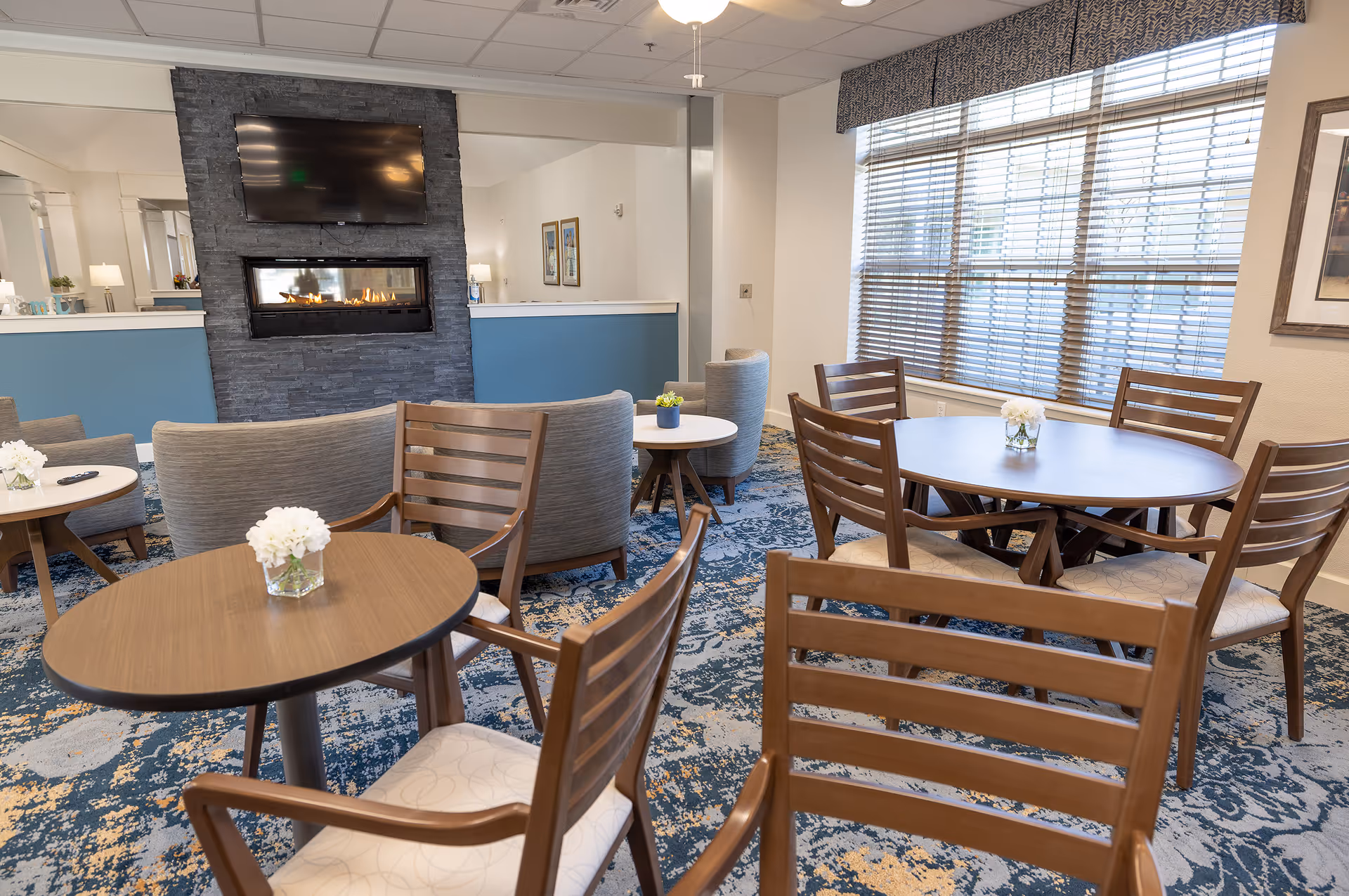 A bright communal dining and lounge area with round wooden tables and chairs, a fireplace with a TV above it, and large windows with blinds.