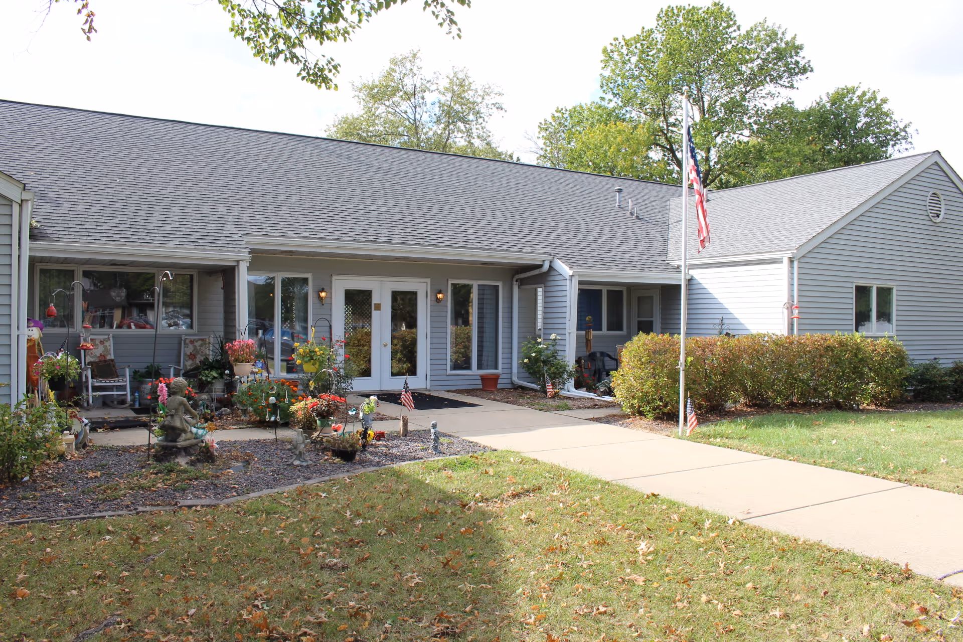 Exterior view of a single-story building with gray siding and a dark shingled roof. There is a concrete walkway leading to double glass doors at the entrance. The area in front of the building features a small garden with various plants, flowers, and decorative items. An American flag is displayed on a flagpole near the entrance. Trees and bushes surround the building.