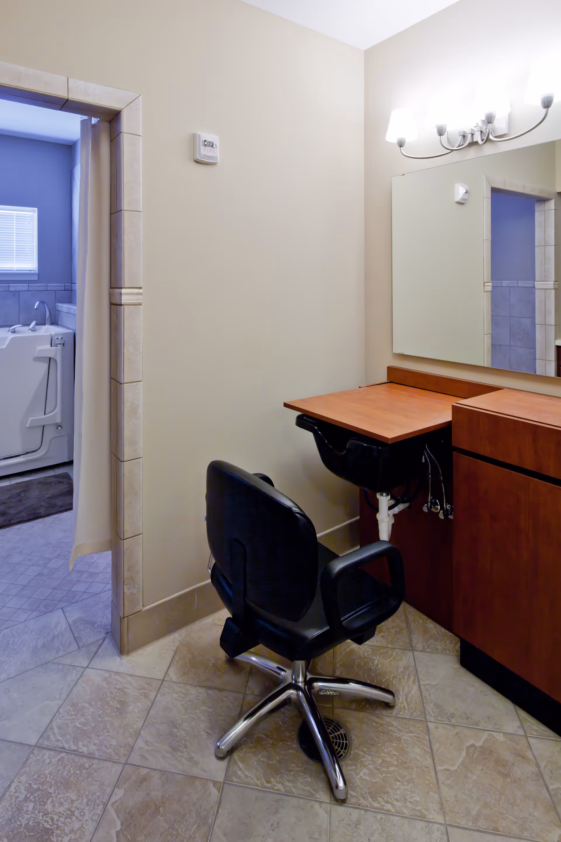 Interior view of a room with a black salon chair in front of a wooden counter with a mirror and light fixture above. To the left, there is an open doorway leading to a bathroom area with a walk-in bathtub and a window with blinds.