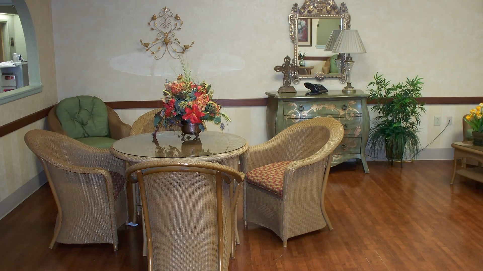 Small common room with a round glass-top table surrounded by wicker chairs, a floral centerpiece, decorative dresser with lamp and mirror, and potted plants.