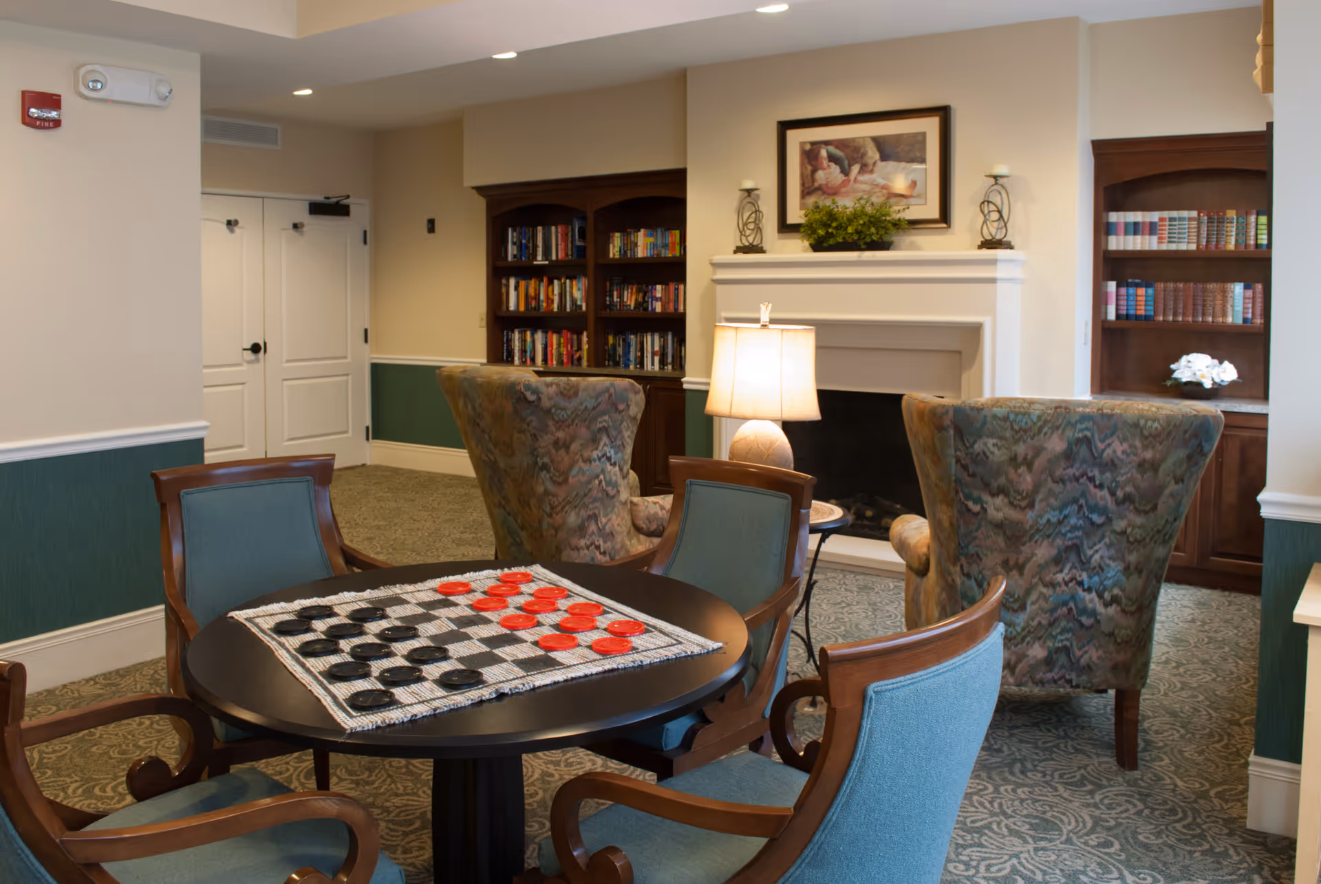 A cozy assisted-living common room with a table set up for checkers, upholstered chairs, bookshelves, and a fireplace.