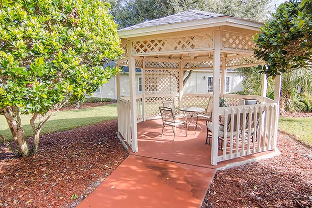 A white wooden gazebo with a table and chairs sits beside a red pathway amid mulch and green shrubs.