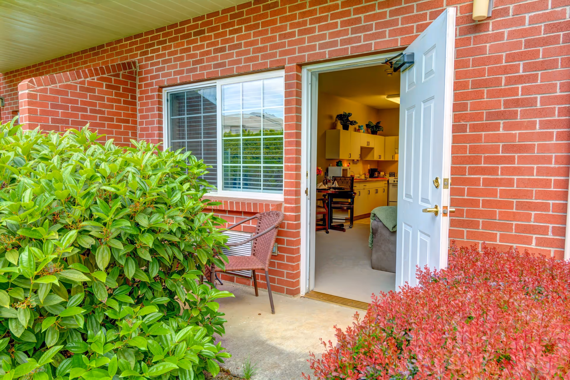 View of a brick building exterior with an open white door leading into a room with kitchen cabinets and a small table with chairs. There are green and red bushes in front of the entrance and a brown chair on the porch.
