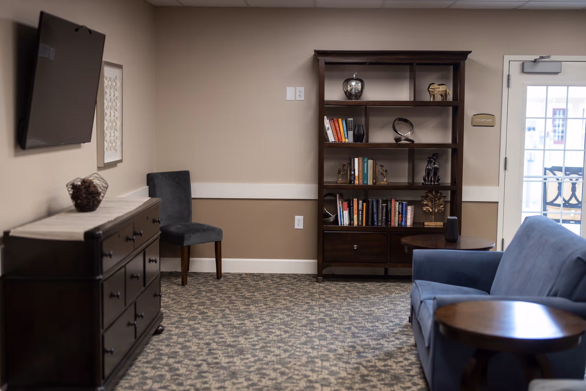 A cozy common room with a blue sofa, wooden bookshelf filled with books and decor, a wall-mounted TV, and a dresser.