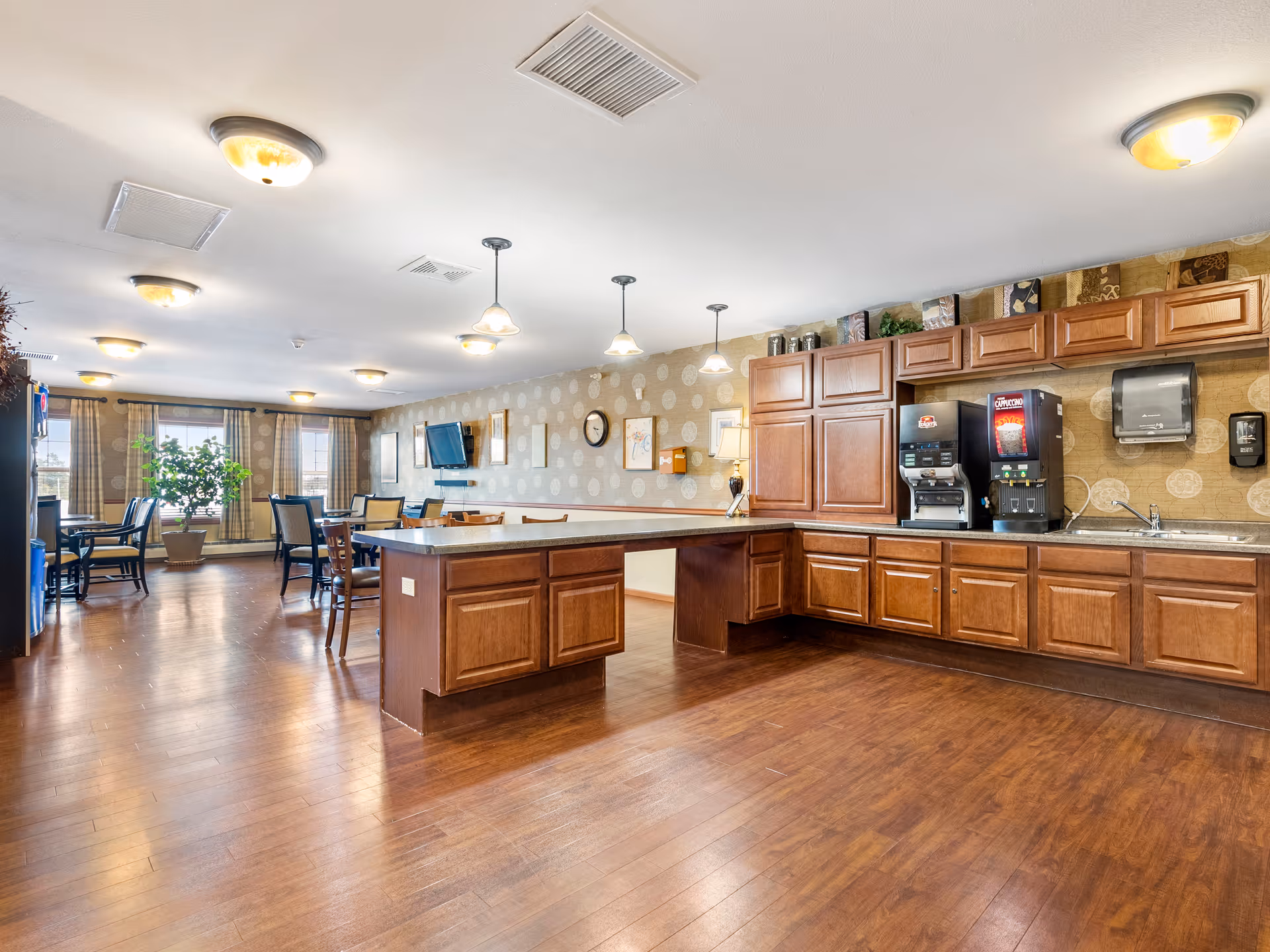 A spacious dining area in a senior living facility featuring wooden floors, multiple tables and chairs, a large kitchen island with cabinets, beverage dispensers, a sink, and wall-mounted television. The room is well-lit with ceiling lights and pendant lamps, and decorated with patterned wallpaper and framed pictures.