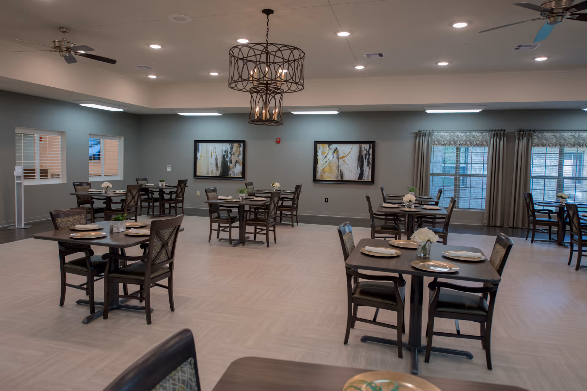 A spacious dining room with multiple dark wood tables and chairs arranged neatly. Each table is set with plates and napkins, and small flower arrangements serve as centerpieces. The room has light-colored flooring, soft blue-gray walls, large windows with curtains, two abstract paintings on the wall, ceiling fans, and a decorative chandelier hanging from the ceiling.