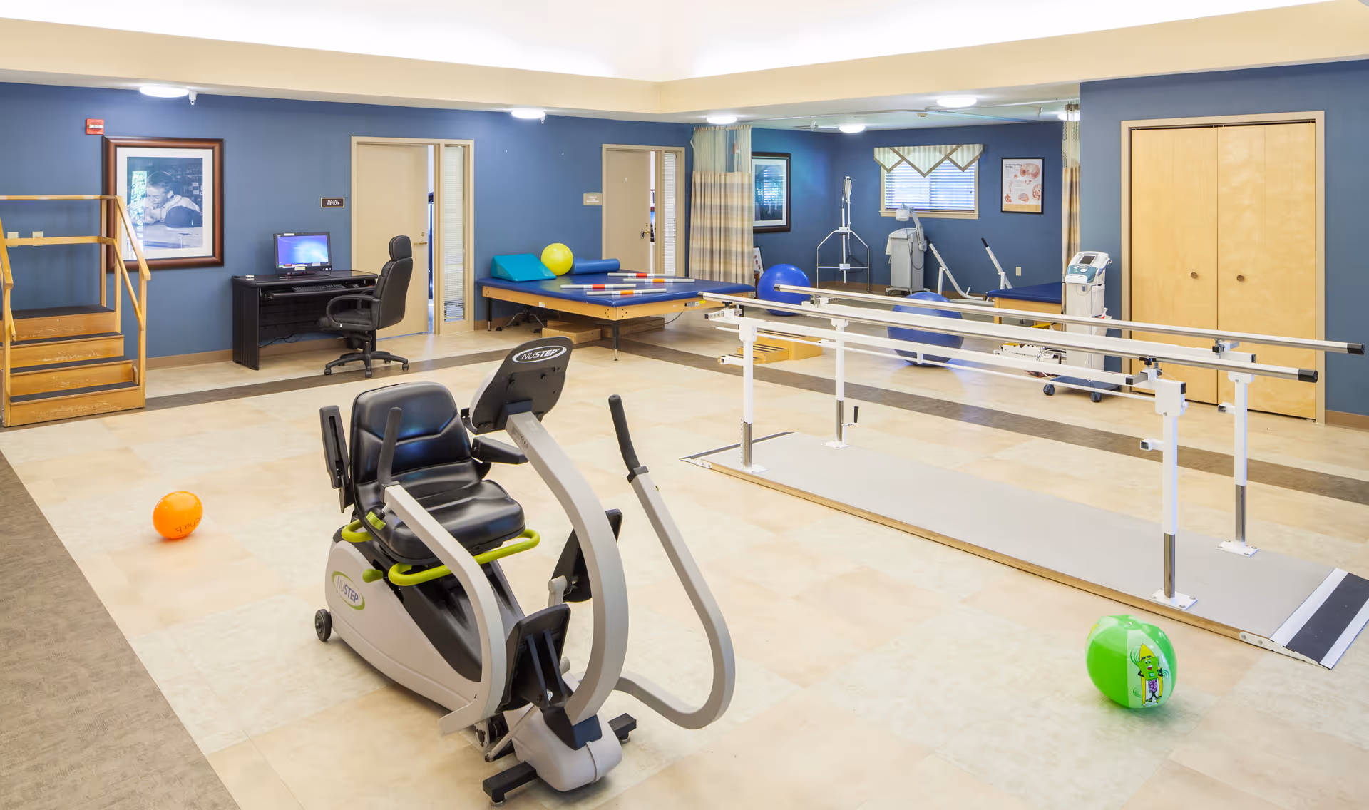 A spacious rehabilitation or physical therapy room with exercise equipment including a recumbent exercise bike, parallel bars for walking practice, therapy balls, and a raised platform with stairs. The room has blue walls, beige flooring, and a desk with a computer in the corner.