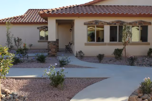 Exterior view of a single-story building with a tiled roof and beige walls, featuring a walkway surrounded by desert landscaping with small bushes and rocks. There are windows and a covered entrance with two chairs visible.