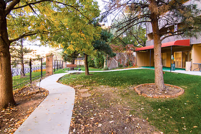 Outdoor garden area with a curved concrete pathway, green grass, several trees with autumn leaves, a bench near a black metal fence, and part of a building with a red awning in the background.
