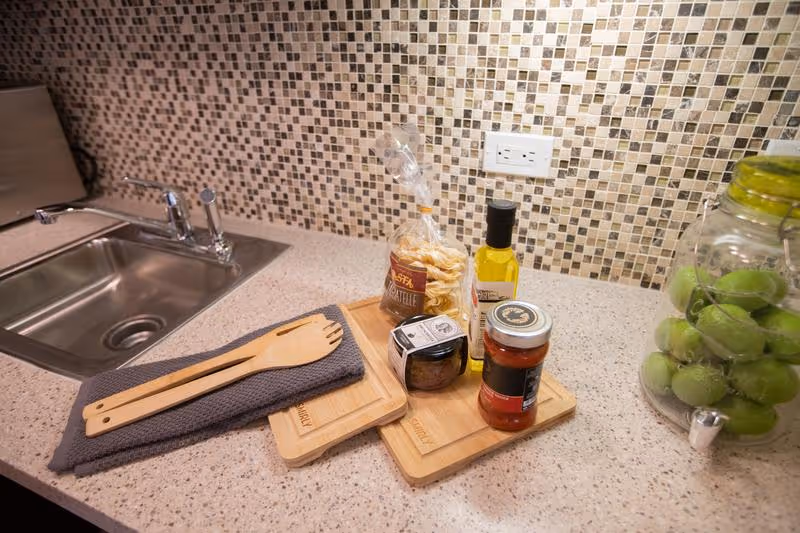 Kitchen countertop with a stainless steel sink on the left, a gray towel with wooden utensils on top, two wooden cutting boards, a bag of pasta, a small bottle of olive oil, a jar of sauce, a small jar of seasoning, and a large glass jar filled with green apples on the right.