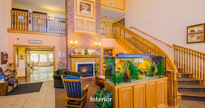 A spacious senior living facility interior with a large fish tank on a wooden stand in the foreground, a cozy seating area with armchairs and a fireplace with brick surround in the background, and a wooden staircase leading to an upper level. The room is warmly lit and decorated with framed artwork and plants.