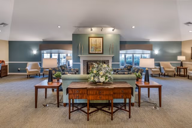 Spacious senior living lounge with sofas and armchairs arranged around a central fireplace, side tables, lamps, and a floral centerpiece on a console table.