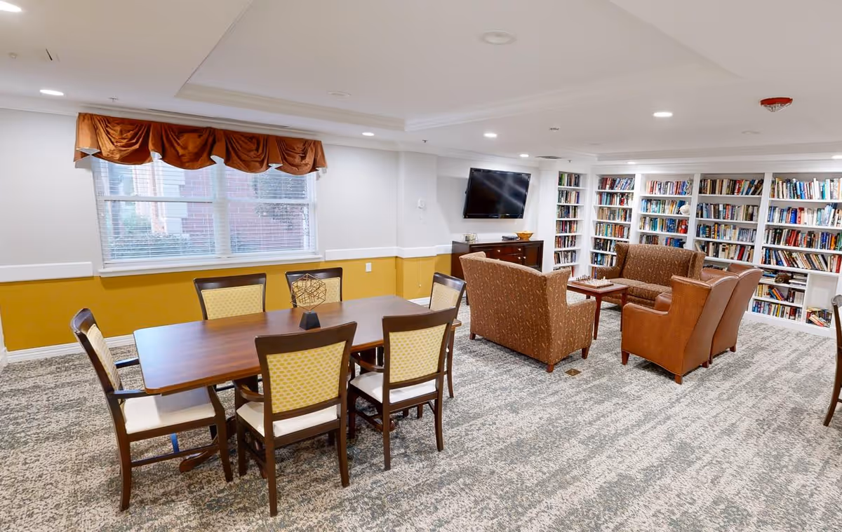 A well-lit room with a large wooden table surrounded by six chairs with yellow patterned cushions. In the background, there is a seating area with two brown armchairs and a brown loveseat facing a wall-mounted flat-screen TV. The walls have built-in bookshelves filled with books, and a window with a brown valance is visible on the left side. The room has a carpeted floor and recessed ceiling lights.