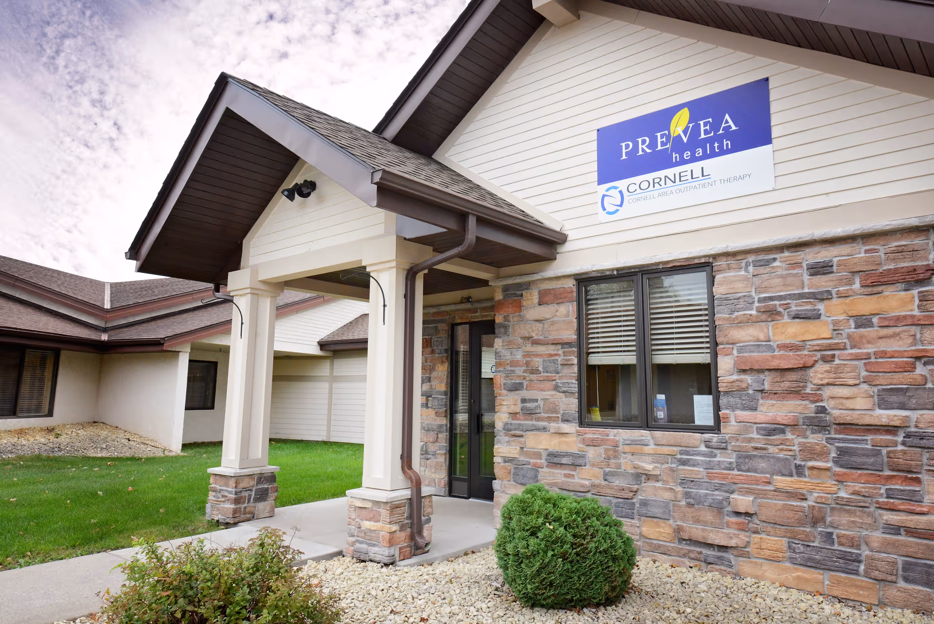 Exterior view of a building entrance with a covered porch supported by columns. The building has stone and siding walls, a window with blinds, and a sign above the entrance that reads 'Prevea Health Cornell Area Outpatient Therapy'. There is a small bush and landscaping with rocks and grass around the entrance.