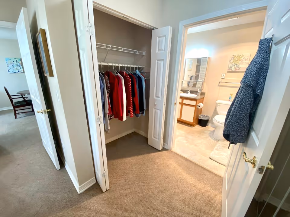 View of a hallway in a senior living facility showing an open closet with clothes hanging on a rod, a bathroom with a toilet, sink, and mirror, and a glimpse of a dining area with a table and chairs in the background.