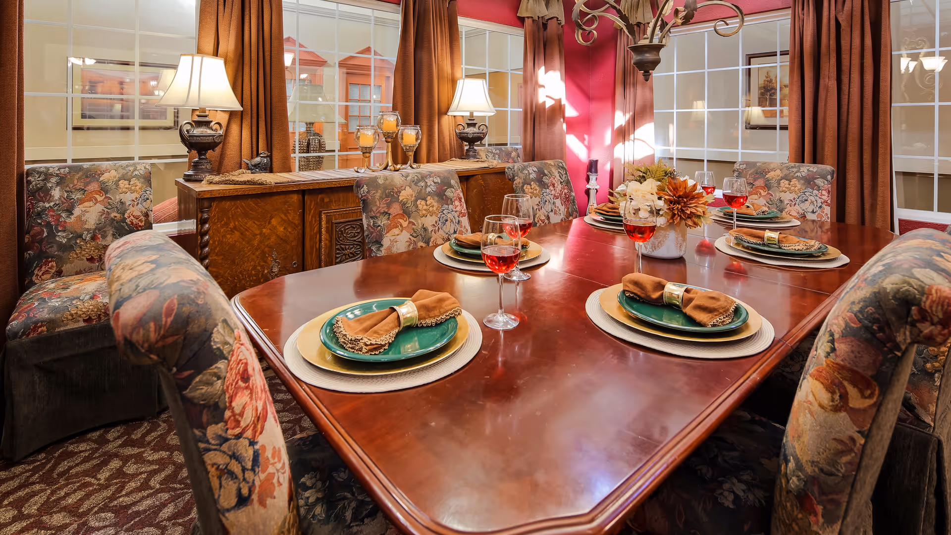 A warmly lit dining room with a polished wooden table set for six, floral-upholstered chairs, wine glasses, and a centerpiece.