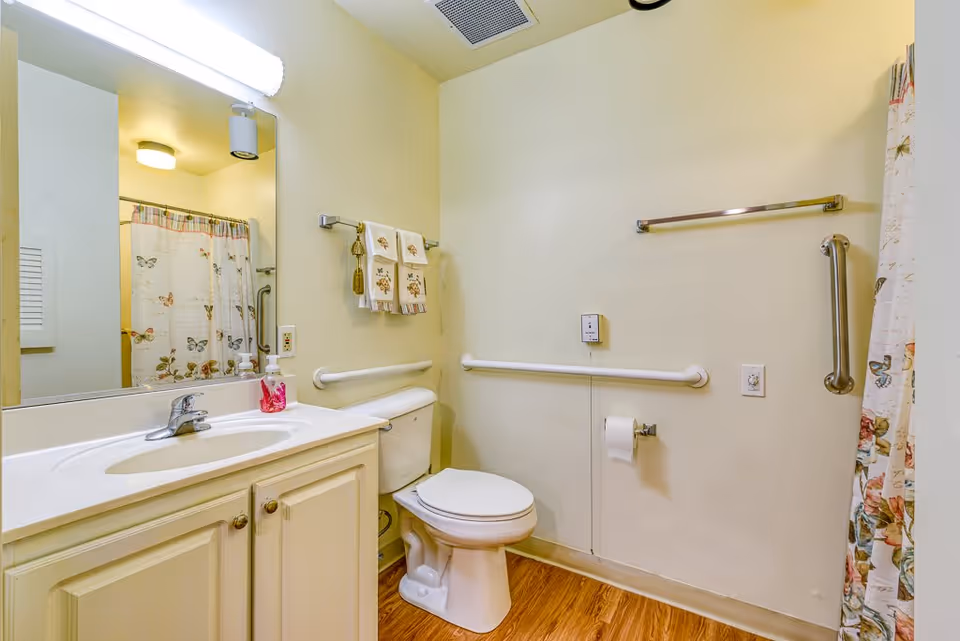 A bathroom with a white sink and cabinet, a toilet, and a shower with a floral and butterfly patterned curtain. There are safety grab bars on the walls near the toilet and shower. A towel rack with folded towels is mounted on the wall above the toilet. The floor has a wood-like finish.