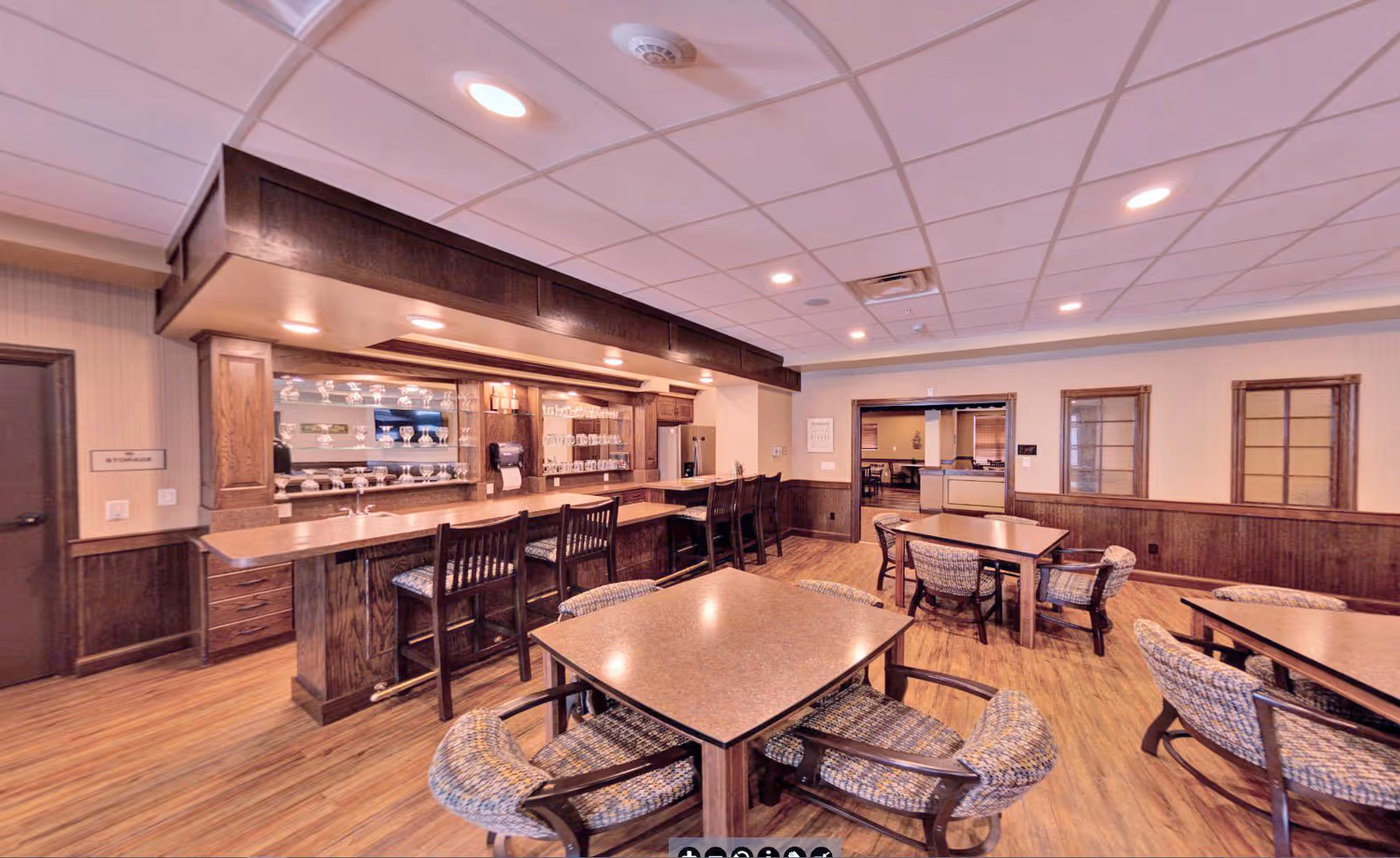 Community dining area with a wooden bar and high stools, several tables and upholstered chairs under recessed ceiling lights.