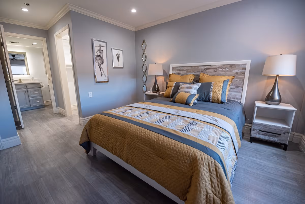 A neatly made bed with a wooden headboard and a quilted bedspread in shades of gold, blue, and gray in a bedroom. Two bedside tables with modern lamps flank the bed. The room has light gray walls and flooring, with two framed fashion sketches on the wall. An open doorway leads to a bathroom with a vanity and mirror visible.