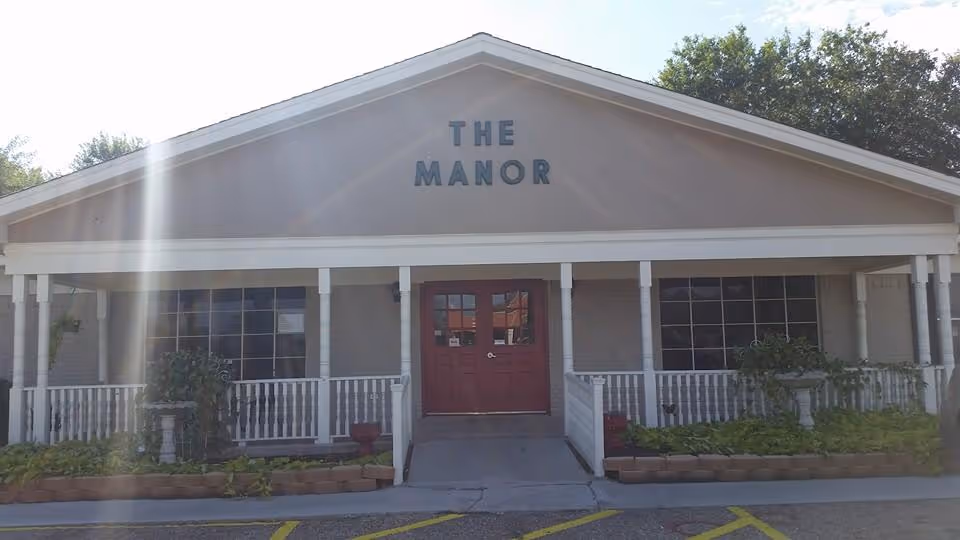 Front exterior view of The Manor Healthcare Residence building with a peaked roof, red double doors, white railings, and plants along the front porch.