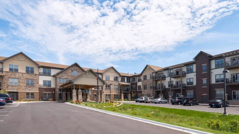 Exterior view of a three-story retirement living facility with a covered entrance, multiple windows, balconies, parked cars, and a partly cloudy sky.