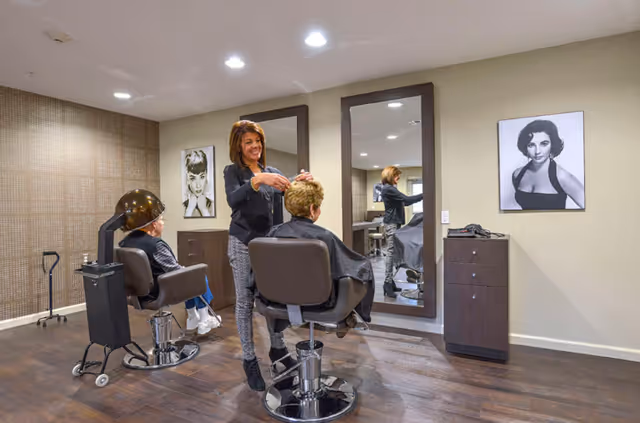 A hair salon area inside a senior living facility with a hairstylist cutting a seated woman's hair. Another woman is seated under a hair dryer. The room has wooden flooring, two large mirrors on the wall, and black-and-white portraits of women on the walls.