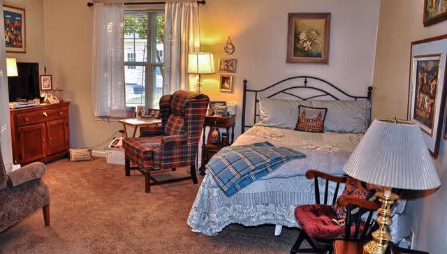 A cozy bedroom in a memory care facility featuring a bed with a decorative pillow and a blue plaid blanket, a plaid armchair next to a window with white curtains, a wooden nightstand with a lamp and framed photos, a wooden dresser with a TV, and various framed pictures on the walls.