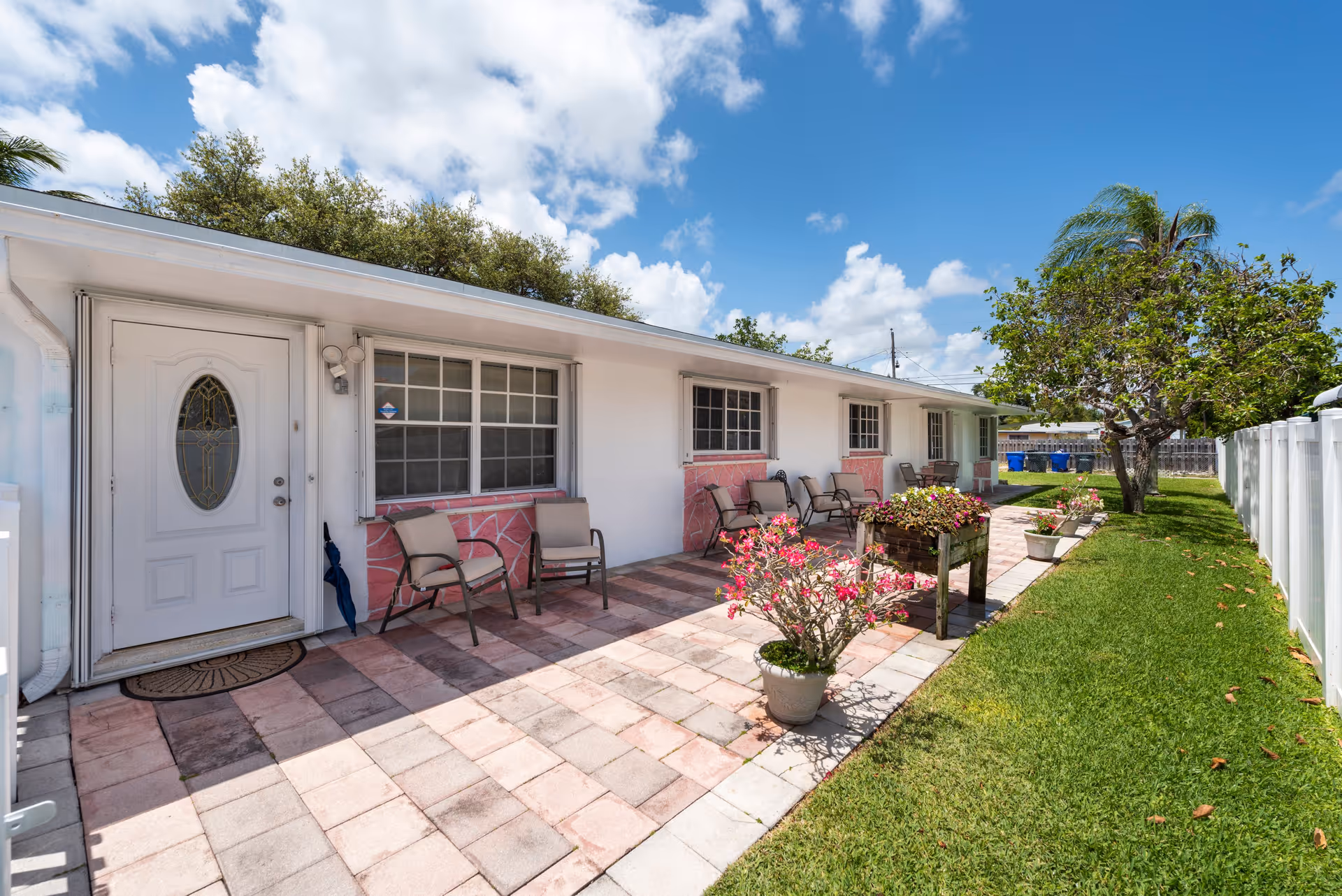 Front exterior of a single-story assisted living building with a tiled patio, outdoor chairs, potted flowers, and a grassy yard under a blue sky.