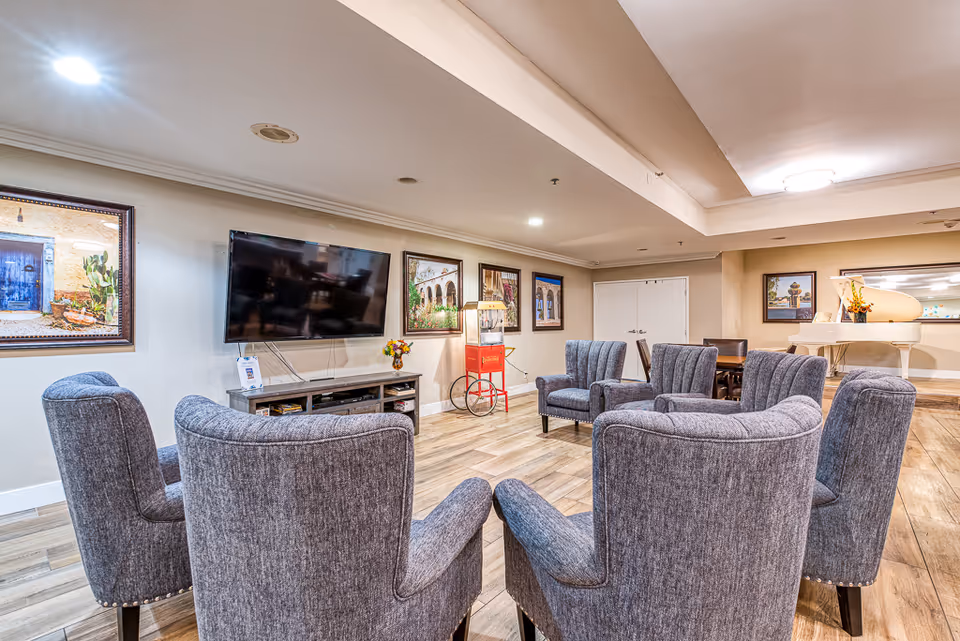 A furnished communal lounge with gray upholstered chairs arranged in a circle facing a wall-mounted TV, wood flooring, and framed artwork.