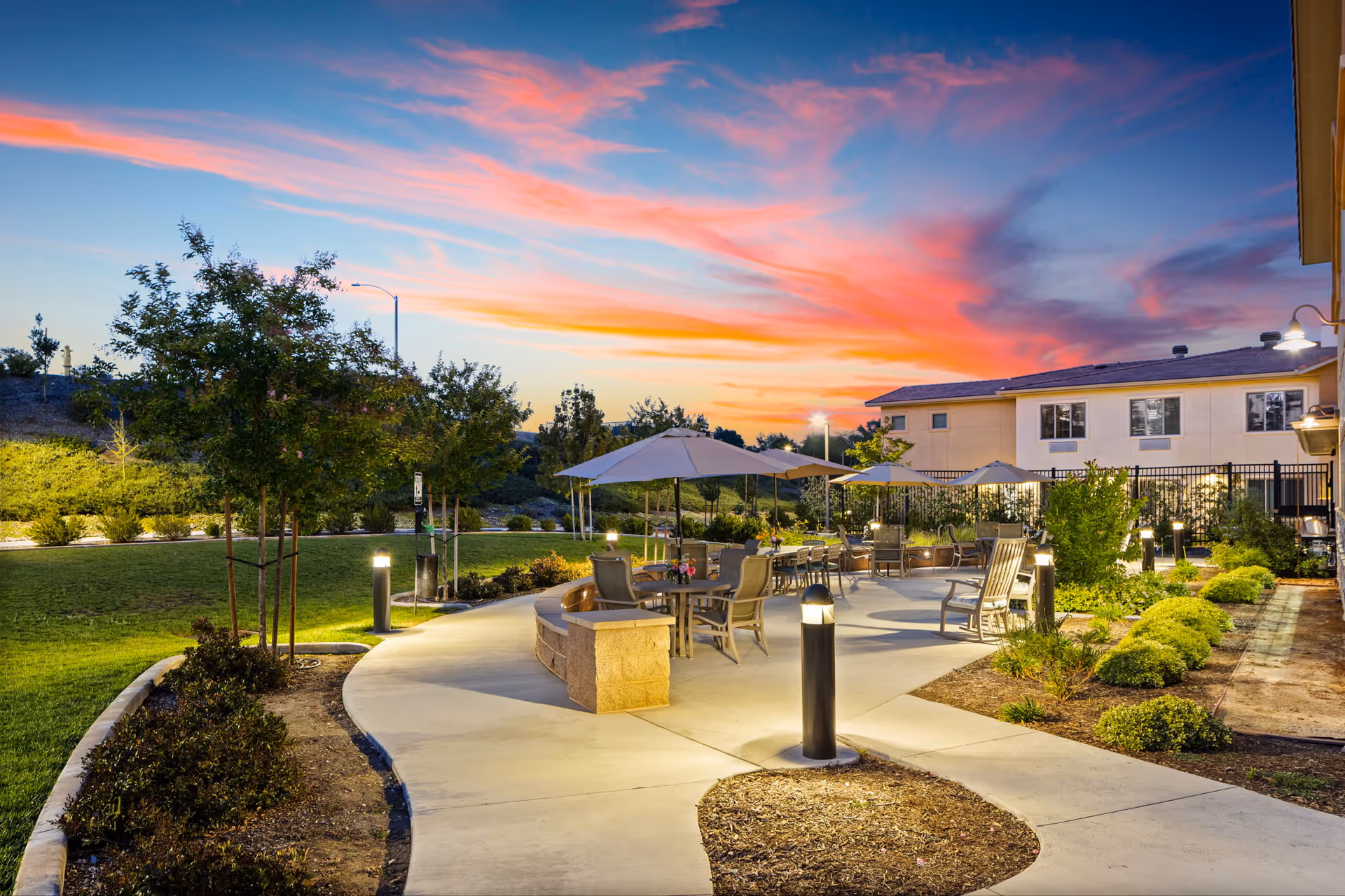 Well-lit outdoor courtyard with tables, umbrellas, chairs, and walkways beside a senior living building at sunset.