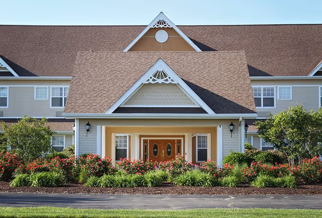 Front exterior view of a senior living facility with a peaked roof, decorative trim, double wooden doors, and windows on either side. The entrance is surrounded by well-maintained landscaping with green bushes and red flowers.