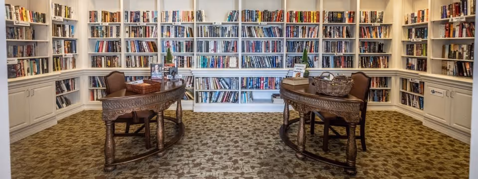 A cozy library room with white built-in bookshelves filled with books lining the walls. Two ornate wooden desks with chairs are positioned facing each other in the center of the room, each desk holding baskets and small decorative items. The floor is covered with a patterned carpet.