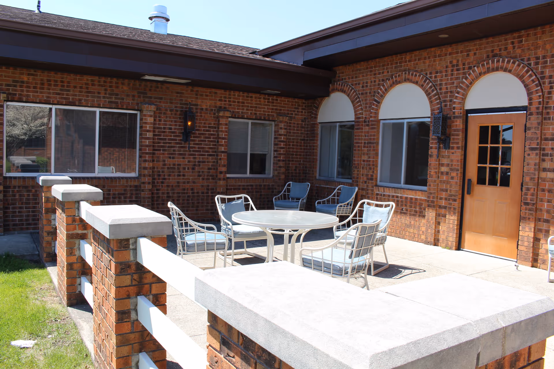 Outdoor patio area with a round glass table surrounded by six metal chairs with cushions. The patio is enclosed by a low brick wall with white railings. The building exterior is made of red brick with several windows and a wooden door with glass panes. The sky is clear and blue.