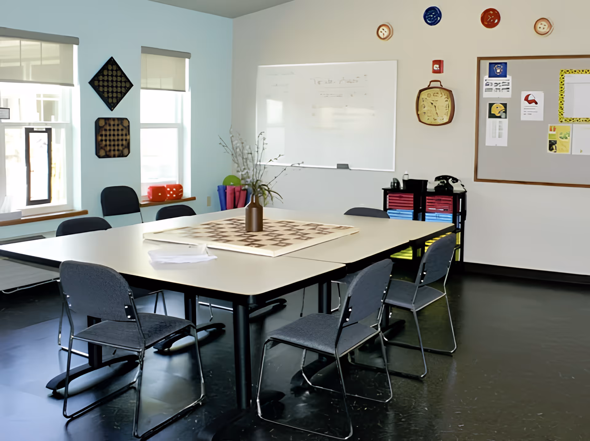 A well-lit room with a large rectangular table in the center surrounded by six chairs. On the table is a vase with branches and a board game. The room has two windows with blinds, a whiteboard on the wall, and a bulletin board with various papers and decorations. The floor is dark and shiny.