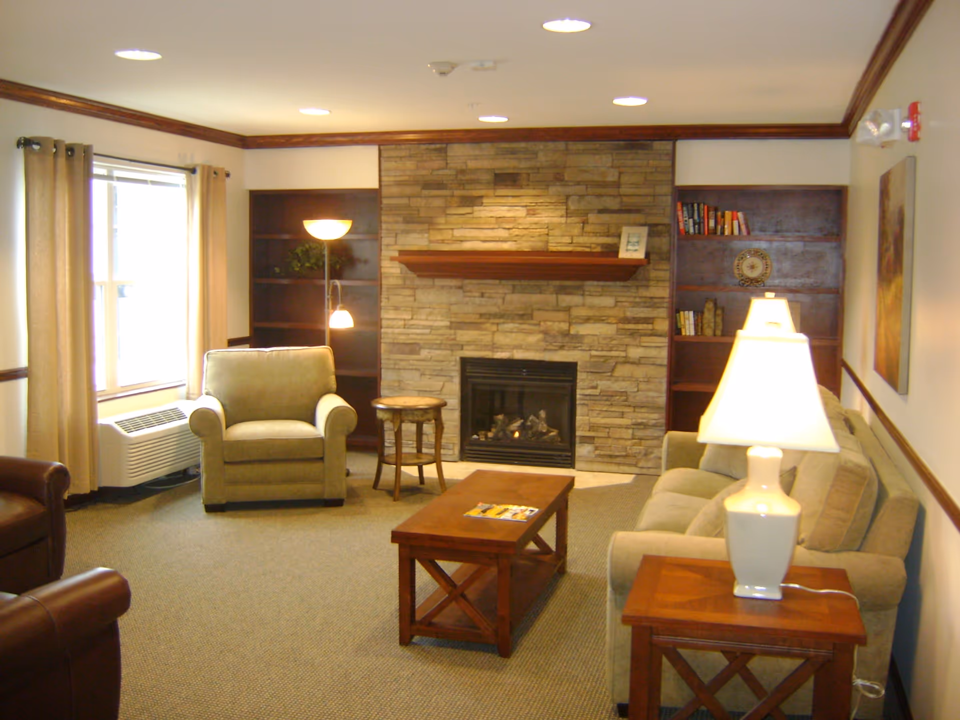 A cozy living room with a stone fireplace centered on the back wall, flanked by dark wooden built-in bookshelves. The room features a beige armchair, a matching sofa, a wooden coffee table, and a side table with a white lamp. There is a window with beige curtains on the left side and recessed ceiling lights illuminating the space.