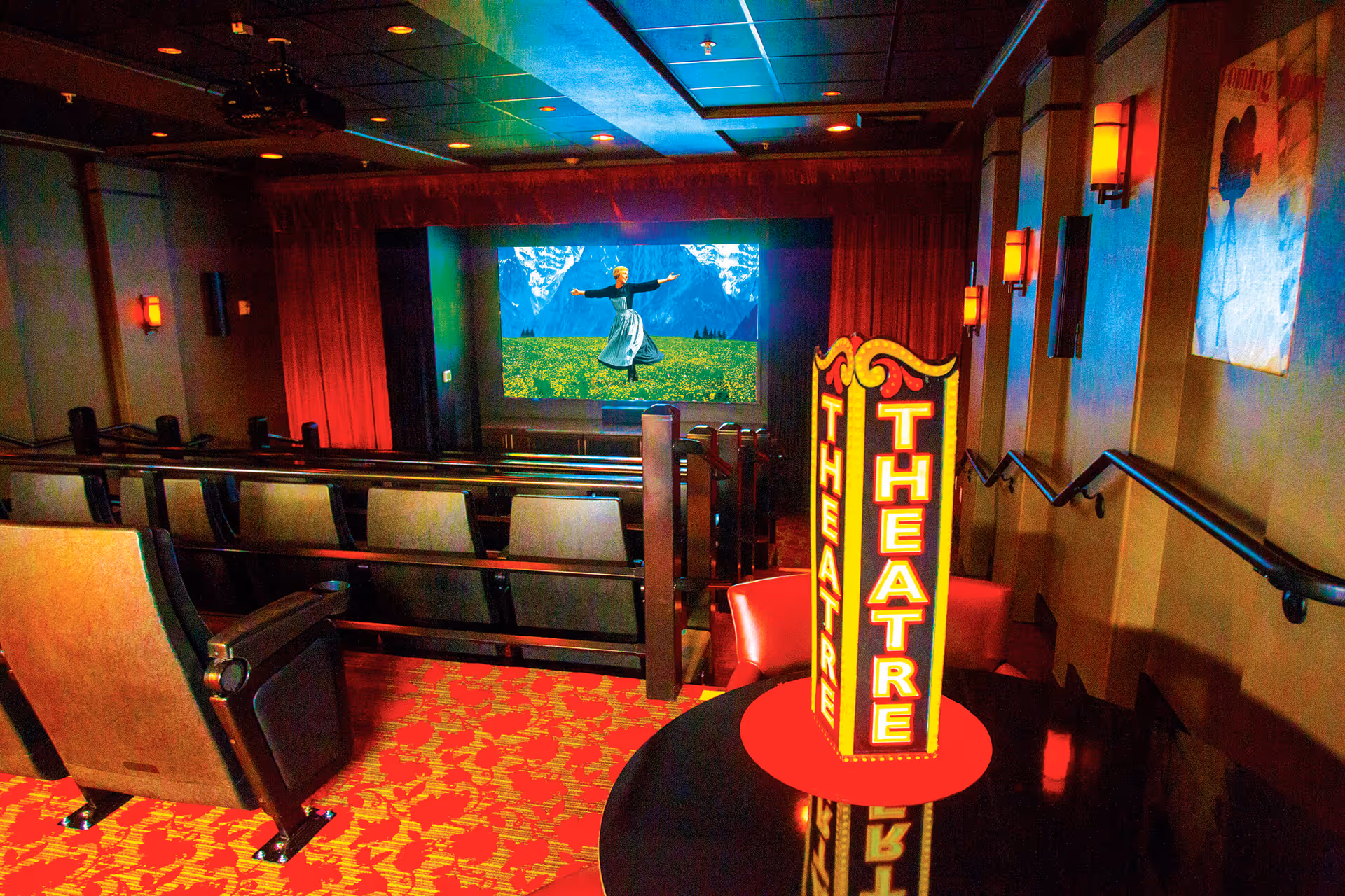 Interior view of a small theater room with rows of cushioned seats facing a large screen showing a scene from The Sound of Music. The room has red patterned carpet, wall sconces with warm lighting, and a decorative illuminated sign reading 'THEATRE' on a round table near the entrance.